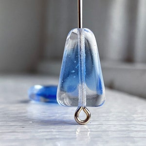 Puede incluir: Una cuenta de vidrio transparente con un tono azul claro y un bucle plateado en la parte inferior. La cuenta está suspendida de un alambre plateado.