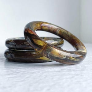 May include: Three stacked bangle bracelets with a brown, black, and white marbled pattern.