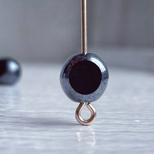 May include: A black hematite bead with a silver loop attached to a silver wire. The bead is round and has a smooth, shiny surface.