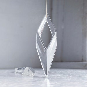 May include: A clear, diamond-shaped crystal ornament hanging from a string. The ornament is suspended above a smaller, clear crystal ornament.