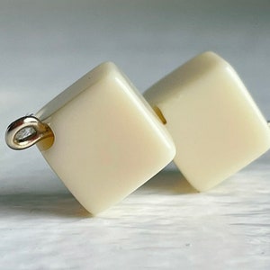 May include: Close-up of two ivory-colored square beads with rounded edges. Each bead has a small silver loop attached, and a hole for stringing. The beads are likely for jewelry making or crafts.