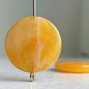 Könnte beinhalten: Zwei runde, gelbe und weiße marmorierte Plastikperlen. Die Perlen liegen auf einer weißen Oberfläche.