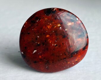 Può includere: Una pietra liscia e rotonda di colore rosso con macchie nere e bianche. La pietra è lucidata e ha una finitura lucida.