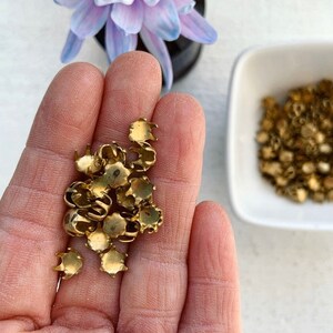 May include: A close-up of a hand holding a pile of small, gold-colored metal settings. The settings have a rounded, cup-like shape with a raised edge. They are likely used for setting gemstones or other decorative elements.