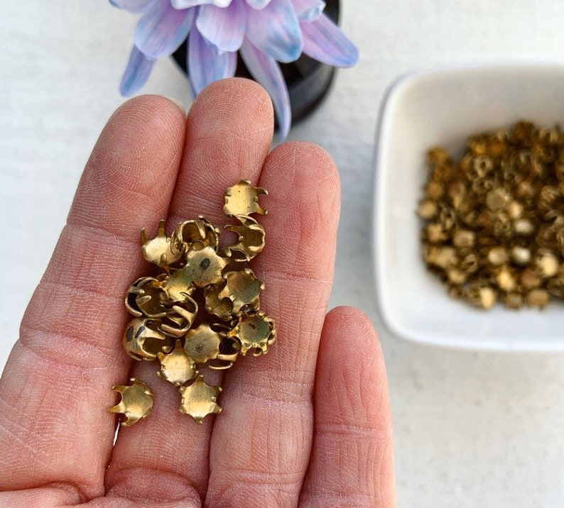 May include: A close-up of a hand holding a pile of small, gold-colored metal settings. The settings have a rounded, cup-like shape with a raised edge. They are likely used for setting gemstones or other decorative elements.