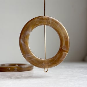 May include: Two large, round, brown resin beads with a marbled white pattern are suspended on a gold-colored wire. The beads have a circular shape with a hole in the center, and a smaller bead is visible in the background.