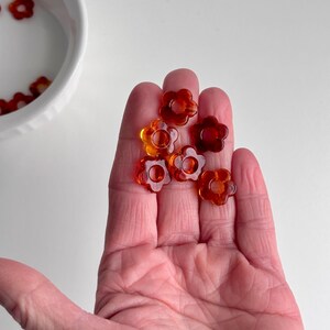 May include: A hand holding several translucent orange flower-shaped beads. The beads have a glossy finish and are scattered across the palm. A white bowl with more beads is in the background.