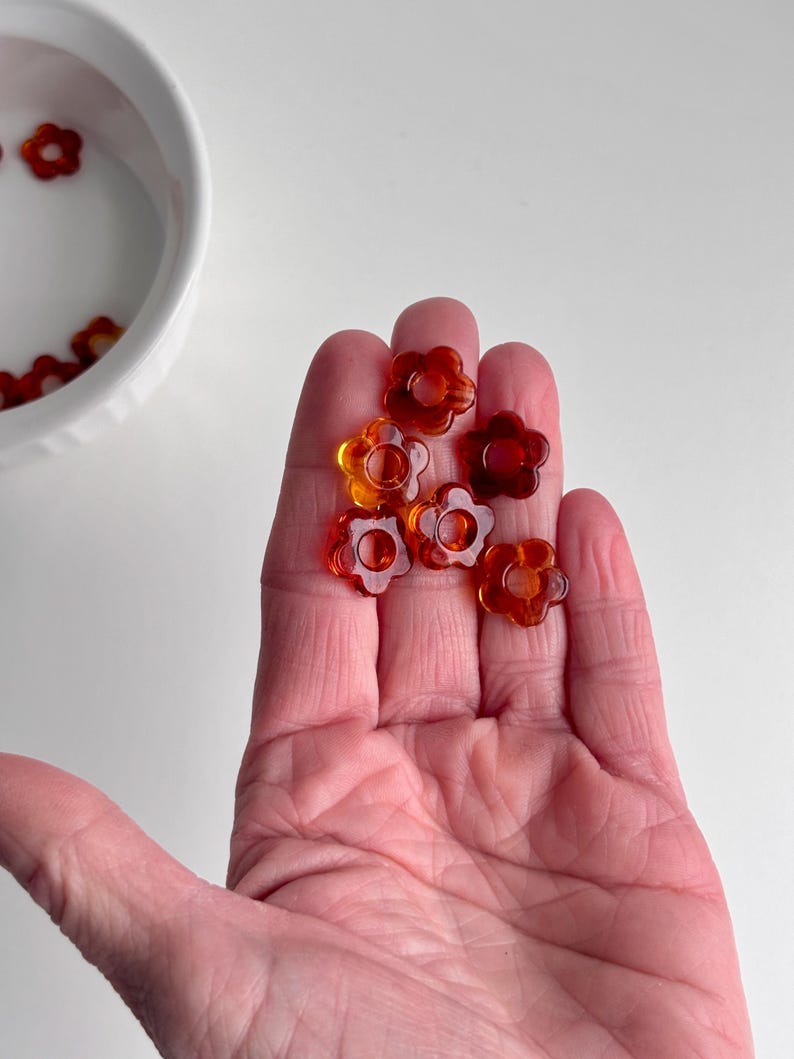 May include: A hand holding several translucent orange flower-shaped beads. The beads have a glossy finish and are scattered across the palm. A white bowl with more beads is in the background.