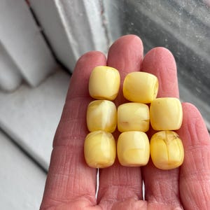 May include: A hand holds nine light yellow, rounded rectangular beads. The beads have a smooth, polished appearance and vary slightly in color, with some showing subtle marbling. The beads are likely for jewelry making or crafting.