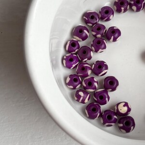 May include: A collection of purple beads with white patterned designs, displayed in a white ceramic bowl. The beads are round with a hole through the center, and the white patterns create a contrasting visual effect.