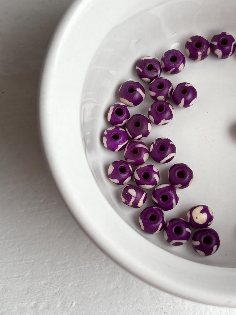 May include: A collection of purple beads with white patterned designs, displayed in a white ceramic bowl. The beads are round with a hole through the center, and the white patterns create a contrasting visual effect.