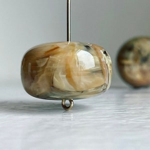May include: A close-up of a light brown and white marbled glass bead with a gold metal loop attached to the bottom. The bead is suspended from a thin metal rod.