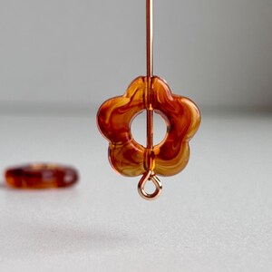 May include: Close-up of a flower-shaped bead with a translucent amber and orange swirl pattern. The bead is suspended on a gold-colored wire, with a smaller, similar bead visible in the background. The design is suitable for jewelry making.
