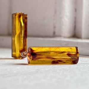 Pode incluir: Duas contas cilíndricas âmbar com efeito marmorizado. Uma conta está em pé, enquanto a outra está na horizontal, presa a um fio fino dourado. As contas têm uma aparência quente e translúcida.