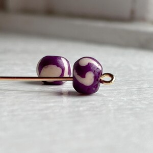 May include: Two purple and cream-colored beads with a swirl pattern are strung on a gold-colored head pin. The beads are round and have a glossy finish. The background is a light, neutral color.