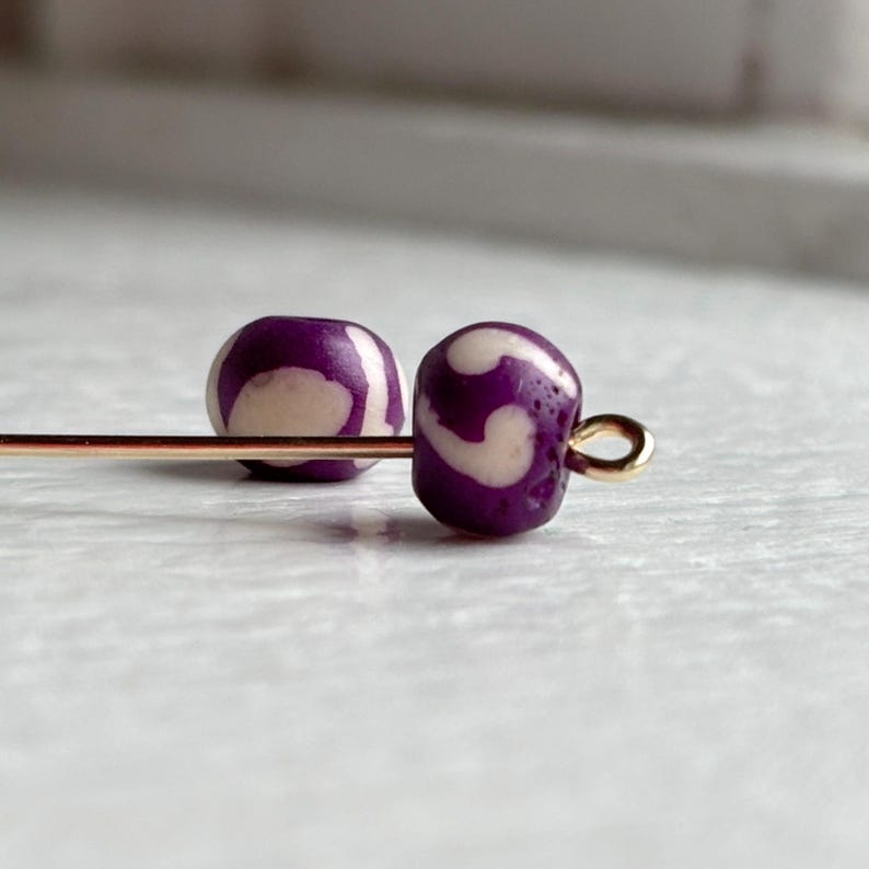 May include: Two purple and cream-colored beads with a swirl pattern are strung on a gold-colored head pin. The beads are round and have a glossy finish. The background is a light, neutral color.