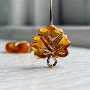 Puede incluir: Un par de pendientes de vidrio ámbar en forma de hoja con un bucle de metal dorado. Los pendientes cuelgan de una superficie blanca.