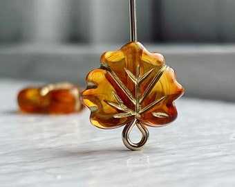 Puede incluir: Un par de pendientes de vidrio ámbar en forma de hoja con un bucle de metal dorado. Los pendientes cuelgan de una superficie blanca.