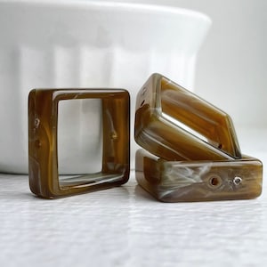 May include: Three brown and white square-shaped plastic frames with rounded edges. The frames have a swirl pattern and are stacked on top of each other.