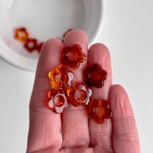 May include: A collection of translucent, amber-colored flower-shaped beads. The beads are held in a hand, with some scattered in a white bowl. The beads have a hole in the center and are a mix of orange and brown hues.