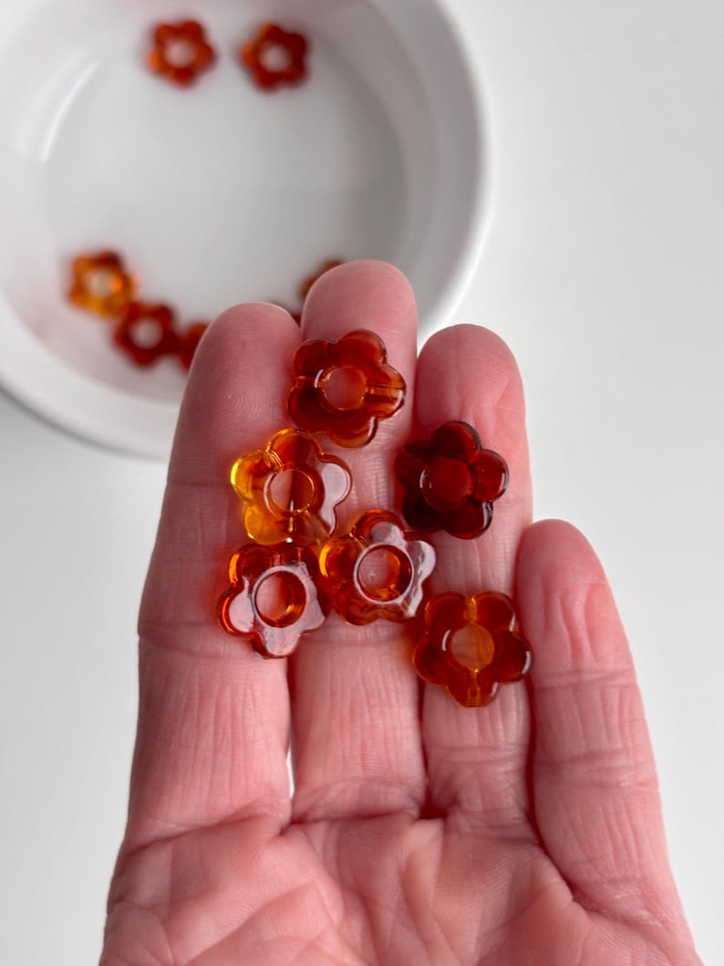 May include: A collection of translucent, amber-colored flower-shaped beads. The beads are held in a hand, with some scattered in a white bowl. The beads have a hole in the center and are a mix of orange and brown hues.