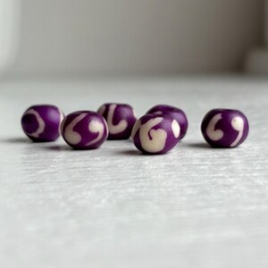 May include: Six purple beads with a white swirl design. The beads are round and have a glossy finish. The beads are arranged on a white surface.