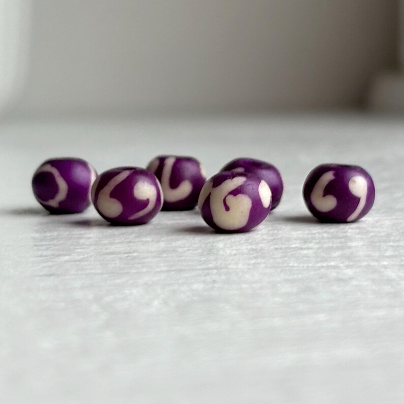 May include: Six purple beads with a white swirl design. The beads are round and have a glossy finish. The beads are arranged on a white surface.