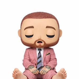 Op de afbeelding: Een vinyl figuur van een man in een roze pak, zittend met zijn benen uitgestrekt. De figuur heeft gesloten ogen, een baard en tatoeages op zijn handen. Hij draagt een gestreepte stropdas en een wit overhemd.