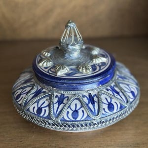 May include: A round ceramic lidded jar featuring a white base with blue painted floral patterns. The lid and body are accented with intricate silver-toned metal filigree overlays and a central pointed metal finial handle on the top.