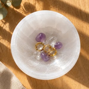 May include: A white, translucent, circular stone bowl containing three gold-toned rings and several small, smooth, polished tumbled stones in shades of purple amethyst and clear quartz, arranged in the center of the bowl.