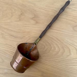 May include: A copper ladle with a dark wooden handle and brass connector. The ladle has a cylindrical bowl with a slightly flared rim. The handle has a small metal ring at the end.