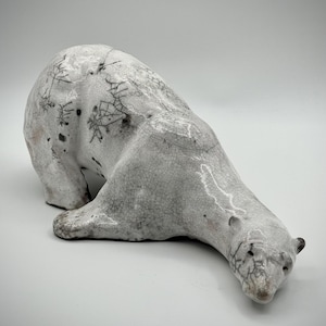 Könnte beinhalten: Eine Keramikskulptur eines Eisbären in liegender Pose. Der Bär ist überwiegend weiß mit einer rissigen, strukturierten Oberfläche und dunklen Markierungen. Der Kopf des Bären ist gesenkt, und seine kleinen Ohren sind sichtbar.