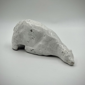 Op de afbeelding: Een keramieken ijsbeerfiguur in liggende houding. De beer is wit met een craquelé-afwerking en kleine donkere vlekken. De sculptuur heeft een gladde, afgeronde vorm, met een langgerekte kop en lichaam.