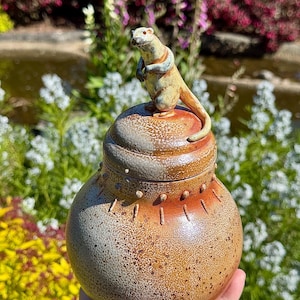 Pode incluir: Um pote de cerâmica artesanal com tampa, apresentando uma figura detalhada de lontra no topo. O pote tem uma paleta de cores terrosas salpicadas de marrom, laranja e cinza. A lontra é marrom claro com detalhes em azul.