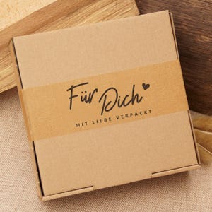 Può includere: Una scatola di cartone marrone con un'etichetta di nastro di carta marrone che dice "Für Dich" con un simbolo di cuore e "MIT LIEBE VERPACKT".