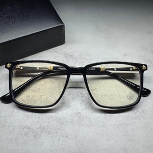 May include: Black rectangular eyeglasses with gold-tone metal accents on the temples and hinges. The frames feature a small fleur-de-lis emblem on the right lens and the text CH8050 printed on the inner temple arm, paired with a black storage box.