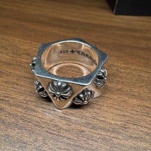 以下が含まれることがあります： 幾何学的なデザインと花のアクセントが特徴のシルバーリング。リングは角が丸い正方形で、".925"の文字とその他の文字が刻まれています。リングは木製の表面に表示されています。