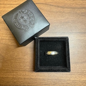 May include: A silver ring with a gold-colored center stone rests in a black velvet-lined box. The ring is displayed in a square black box. A black box with a silver emblem is in the background.