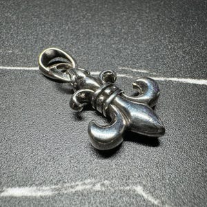 May include: A polished silver-toned metal pendant shaped like a fleur-de-lis. The pendant features a smooth, curved design with a central vertical element and three distinct petal-like sections. It includes a rounded bail at the top for attachment.