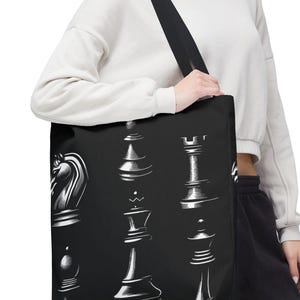May include: Black tote bag featuring a white chess piece design. The bag displays various chess pieces, including a knight, bishop, rook, king, and queen. The bag has a black shoulder strap and is held by a person.