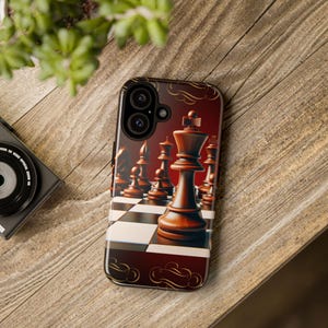 May include: A smartphone case featuring a chess board design with a king piece in the foreground. The case has a glossy finish with a dark red background and gold decorative elements. The phone case is on a wooden surface.