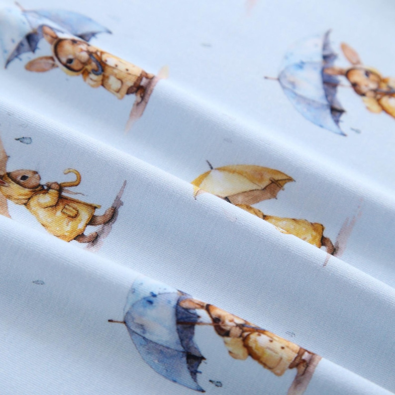 Puede incluir: Tela azul claro con un patr&oacute;n repetido de conejitos de acuarela sosteniendo paraguas. Los conejitos visten impermeables amarillos y marrones, y los paraguas son azules y amarillos. El dise&ntilde;o tiene un aire caprichoso y de cuento.