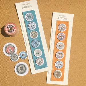 以下が含まれることがあります： 「Porcelain Buttons」というテキストが書かれた2枚のカードと、ヴィンテージデザインのさまざまな磁器ボタン。ボタンは、青とオレンジの生地見本に飾られています。