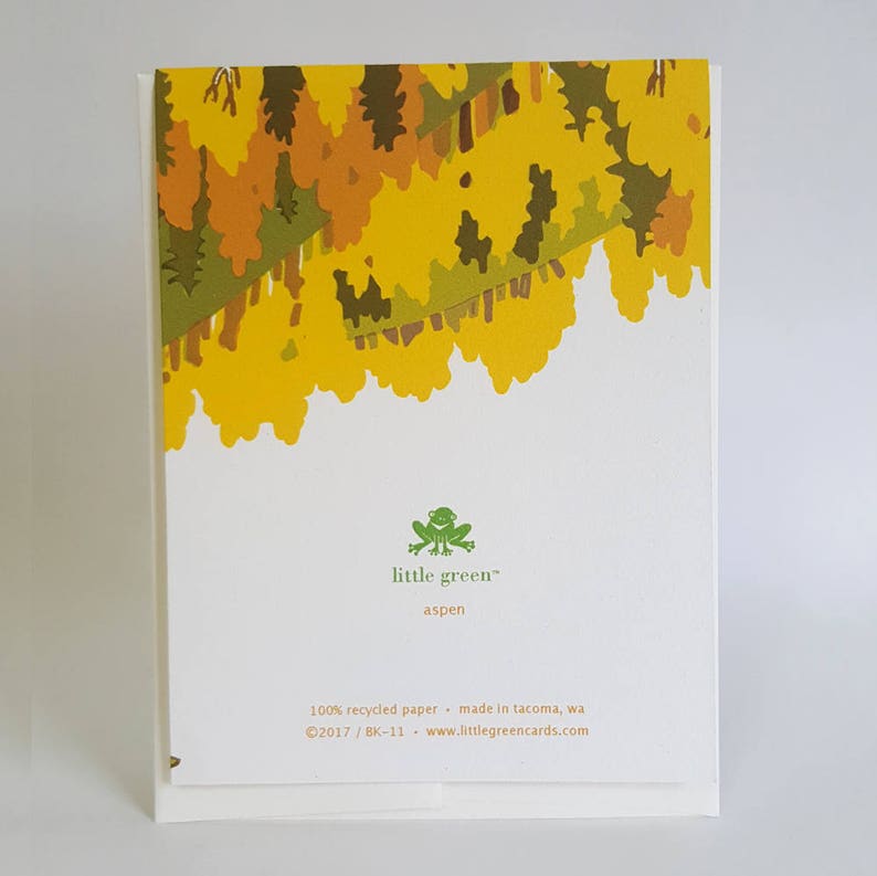 May include: A folded greeting card with a white background and a yellow, brown, and green tree design at the top. The card features the text "little green" and "aspen", along with additional text about the paper and the company.