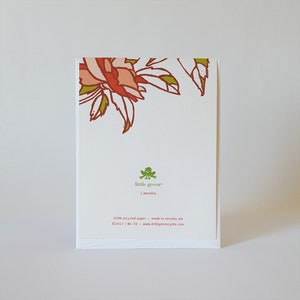May include: Back of a white card with a green frog logo that says "little green" and "Camellia". The card also says "100% recycled paper - made in Tacoma, WA &copy;2017/BK-03 www.littlegreencards.com". The top of the card has a red and pink floral design.