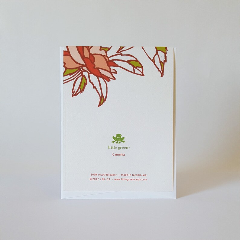 May include: Back of a white card with a green frog logo that says "little green" and "Camellia". The card also says "100% recycled paper - made in Tacoma, WA &copy;2017/BK-03 www.littlegreencards.com". The top of the card has a red and pink floral design.