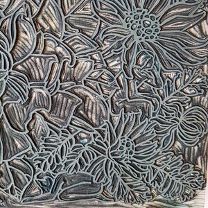 May include: A close-up of a detailed, silver-toned linoleum block print featuring a floral design. The print shows a variety of flowers, including daisies and ferns, with intricate details and textures.