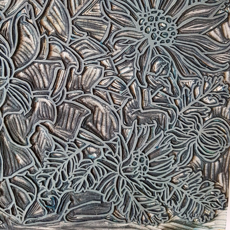 May include: A close-up of a detailed, silver-toned linoleum block print featuring a floral design. The print shows a variety of flowers, including daisies and ferns, with intricate details and textures.