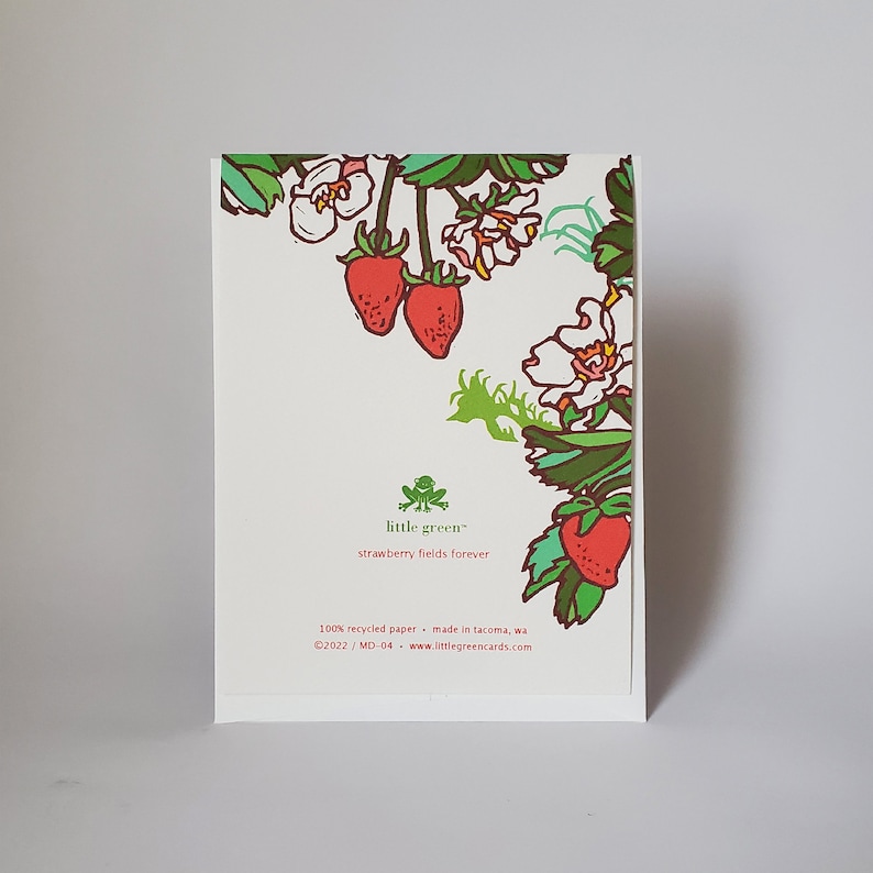 May include: A white card with a green frog logo and the text "little green" and "strawberry fields forever". The card is decorated with a floral design featuring red strawberries and green leaves.