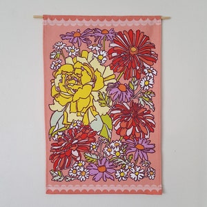 May include: A pink fabric wall tapestry with a floral design. The tapestry features a large yellow rose, red and pink flowers, and white daisies. The tapestry has a scalloped border.